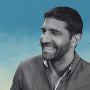 Fotografía en blanco y negro de un hombre con barba corta y cabello oscuro, sonriente y mirando hacia su derecha. Lleva una camisa de botones. El fondo es azul con un ligero degradado hacia amarillo en la parte inferior, simulando un cielo.