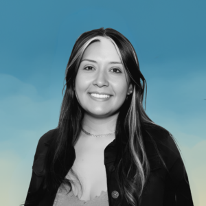 Fotografía en blanco y negro de una mujer joven con cabello largo y oscuro, con mechones claros. Está sonriendo y mirando de frente. Lleva una chaqueta oscura y un collar. El fondo es azul con un degradado hacia amarillo en la parte inferior, simulando un cielo.