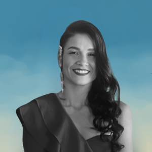 Fotografía en blanco y negro de una mujer joven con cabello largo y oscuro, peinado hacia un lado con ondas. Está sonriendo y lleva un vestido con un hombro descubierto. Usa un pendiente largo en una oreja. El fondo es azul con un degradado hacia amarillo en la parte inferior, simulando un cielo.