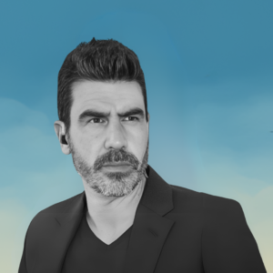 Fotografía en blanco y negro de un hombre con cabello oscuro y barba corta, mirando hacia su izquierda con una expresión seria. Lleva una chaqueta oscura y una camiseta negra. En su oreja tiene un auricular. El fondo es azul con un degradado hacia amarillo en la parte inferior, simulando un cielo.