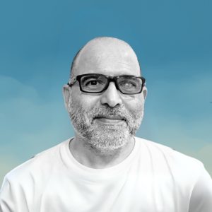 Fotografía en blanco y negro de un hombre calvo con barba canosa y gafas negras, sonriendo suavemente y mirando de frente. Viste una camiseta clara. El fondo es azul con un degradado hacia amarillo en la parte inferior, simulando un cielo.