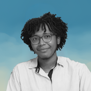 La imagen muestra a un joven de cabello rizado y gafas, mirando directamente a la cámara con una expresión amable. La imagen es en blanco y negro, mientras que el fondo presenta un cielo azul con algunas nubes, creando un contraste entre el retrato y el entorno. Lleva una camisa de rayas claras.