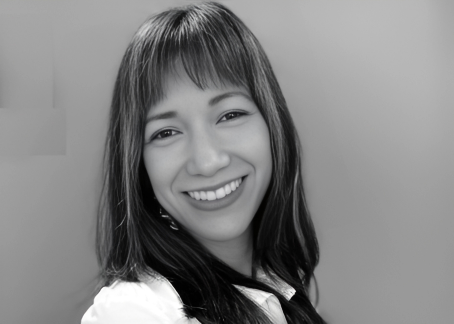 La imagen muestra a una mujer joven sonriendo, con el cabello largo y flequillo. La fotografía es en blanco y negro, y la mujer viste una camisa clara. Su expresión transmite amabilidad y calidez. El fondo es neutro, lo que destaca aún más su rostro sonriente.