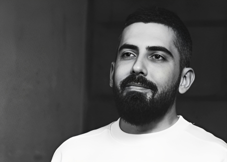 La imagen muestra a un hombre con barba espesa y cabello corto, mirando ligeramente hacia arriba con una expresión tranquila. Viste una camiseta clara y la fotografía es en blanco y negro. El fondo es oscuro y neutro, lo que hace que el rostro del hombre sea el punto focal de la imagen.
