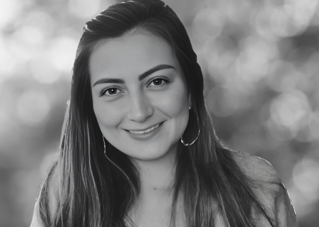 La imagen muestra a una mujer joven sonriendo, con el cabello largo y suelto. Lleva pendientes circulares y viste una prenda clara. La fotografía es en blanco y negro, con un fondo desenfocado que parece representar naturaleza o luces suaves, lo que resalta su expresión amable y relajada.