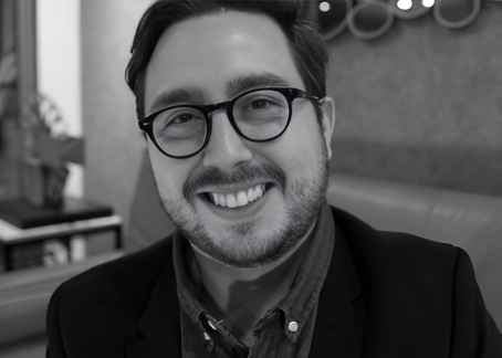La imagen muestra a un hombre joven con gafas, barba corta y una gran sonrisa. Está vestido de manera formal con una chaqueta y camisa. La fotografía es en blanco y negro, y el entorno parece ser un espacio interior acogedor, posiblemente una sala. Su expresión es amigable y relajada, transmitiendo calidez.