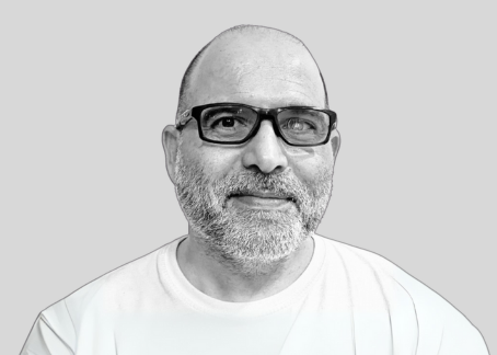 La imagen muestra a un hombre calvo con gafas y barba canosa, sonriendo ligeramente. Lleva una camiseta clara. La fotografía es en blanco y negro con un fondo gris liso, lo que hace que su rostro y expresión sean el enfoque principal. Su expresión transmite calma y amabilidad.