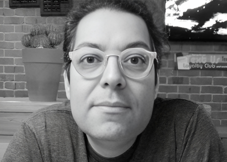 La imagen muestra a un hombre con gafas de montura clara, cabello oscuro y expresión neutral. Está sentado frente a una pared de ladrillo y una planta en una maceta es visible al fondo. La iluminación es suave y parece estar en un ambiente interior, como un café o restaurante.