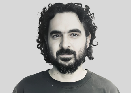 La imagen muestra a un hombre con cabello rizado y barba, mirando directamente a la cámara con una expresión serena. Lleva puesta una camiseta oscura. La fotografía es en blanco y negro, con un fondo claro que resalta el rostro del hombre, destacando sus facciones y mirada calmada.